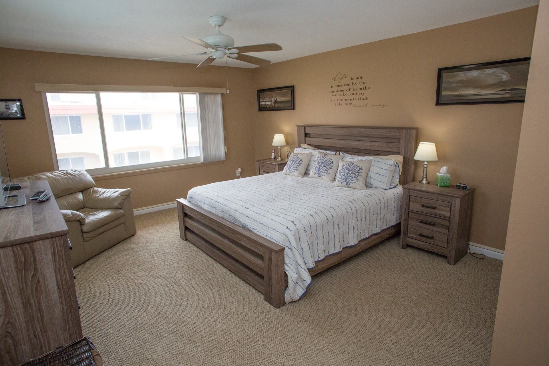 A bedroom with a large bed, two bedside tables with lamps, a desk, a chair, a ceiling fan, and a window with curtains.