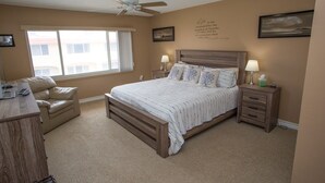 A bedroom with a large bed, two bedside tables with lamps, a desk, a chair, a ceiling fan, and a window with curtains. - Win San A-208 2BD - Sandy Toes & Salty Breezes (New Smyrna Beach)