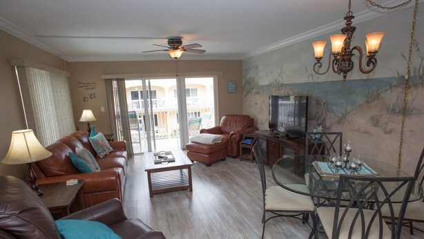 A living room with a brown leather sofa, a glass coffee table, a leather armchair, and a dining area with a glass table and metal chairs. - Win San A-208 2BD - Sandy Toes & Salty Breezes (New Smyrna Beach)