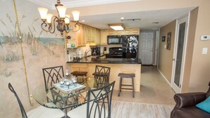 A compact kitchen with a dining area, featuring a glass table, metal chairs, and a brown leather sofa. - Win San A-208 2BD - Sandy Toes & Salty Breezes (New Smyrna Beach)