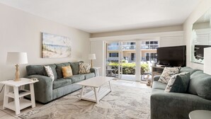 A living room with a grey sofa, a white coffee table, and a TV on a stand. There is a painting on the wall and a balcony with a view of the outside. - Win San C-104 2BD - Beach Life Vibes All Day (New Smyrna Beach)