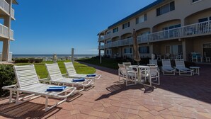 A hotel patio with outdoor seating, a view of the ocean, and a building with multiple balconies. - Win San C-104 2BD - Beach Life Vibes All Day (New Smyrna Beach)