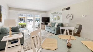 A modern living room with a sofa, a coffee table, a TV, and a dining table with placemats. - Win San C-104 2BD - Beach Life Vibes All Day (New Smyrna Beach)
