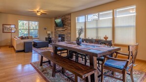 A spacious living room with a large wooden dining table, comfortable seating, a fireplace, and large windows.