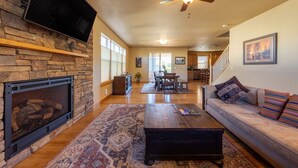 A spacious living room with a stone fireplace, a flat-screen TV mounted on the wall, a ceiling fan, and a large window providing natural light.