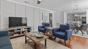 Una sala de estar moderna con un televisor de pantalla plana, sillones azules, una mesa de centro de madera y un ventilador de techo.