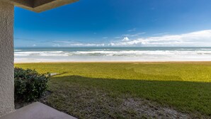 A view from a covered porch overlooking a beach with waves, grassy area, and clear sky. - Errol by the Sea unit 125- 2BD - Beachy Keen (New Smyrna Beach)