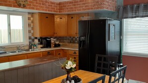 Une cuisine avec des meubles de rangement en bois, un réfrigérateur noir, une table à manger avec des chaises et une fenêtre avec des stores.
