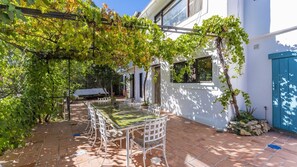 Une terrasse avec une table et des chaises, entourée de verdure et bordée d’un bâtiment blanc.