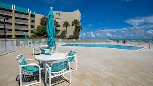 An outdoor pool area with a table and chairs, a turquoise umbrella, and a building with balconies in the background. - Errol by the Sea unit 412- 1BD - Tide Time (New Smyrna Beach)