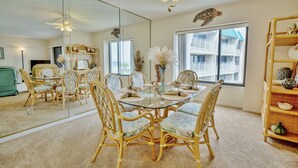 A dining area with a glass table surrounded by wicker chairs, a ceiling fan, and a large window with a view of a building. - Errol by the Sea unit 412- 1BD - Tide Time (New Smyrna Beach)