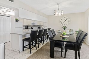 Een moderne keuken met witte kastjes, een magnetron en een koffiezetapparaat. Er staat een eettafel met zwarte stoelen en een klok aan de muur.