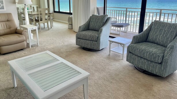 Amplia sala de estar con un gran ventanal que ofrece vistas al océano. Hay dos sillones grises, un sofá beige y una mesa de centro blanca.