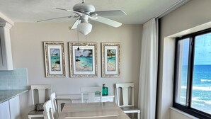 Un comedor con una mesa larga y sillas blancas, un ventilador de techo, cuadros de playas enmarcados, y vista al océano a través de un ventanal grande.