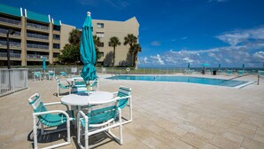 Zona de piscina al aire libre con una mesa y sillas, una sombrilla turquesa y un edificio con balcones al fondo.