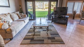 A living room with a sofa, a glass-top coffee table, a TV, and a view of a courtyard with buildings. - Errol by the Sea - Unit 107 - 1BD - The Salty Nest (New Smyrna Beach)