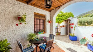 Un patio cubierto con una mesa y sillas, rodeado de plantas en macetas y una pared blanca.