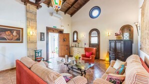 Un vestíbulo amplio con un sillón rojo, un sofá y una mesa de centro de vidrio. Se ve una puerta de madera y una escalera al fondo.