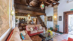 Una sala de estar acogedora con techo alto, vigas de madera y una escalera. Hay un comedor con mesa y sillas, y una cocina al fondo.