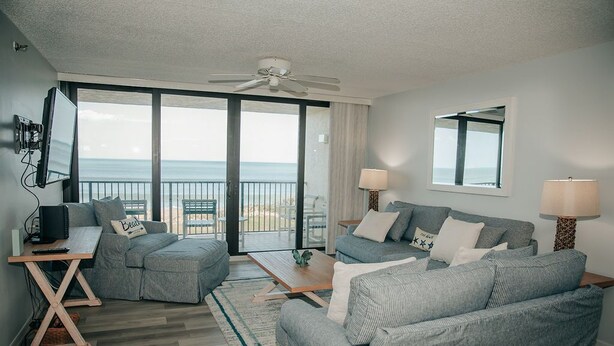A modern living room with a large sliding glass door leading to a balcony, a ceiling fan, and a flat-screen TV on a wooden stand.