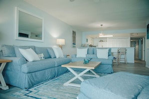 A living room with a sofa set, a coffee table, and a dining area with a kitchen in the background. - Chadham by the Sea unit 213- BD- Beach Bliss (New Smyrna Beach)