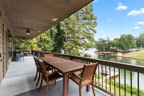 A covered outdoor patio with a wooden table and chairs, overlooking a body of water and greenery.