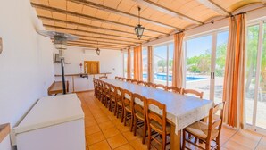 Un comedor con una mesa larga y sillas, un refrigerador, techo de madera y ventanas grandes con cortinas.