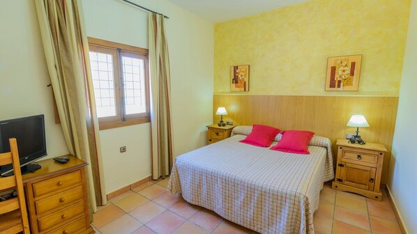 Un dormitorio con una cama, dos mesitas de noche con lámparas, un televisor sobre un aparador de madera y una ventana con cortinas.