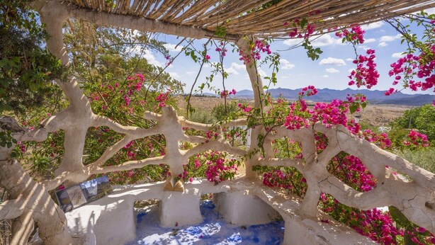 Een wit gebouw van steen, versierd met roze bougainvillea, een houten pergola en een bergachtig landschap op de achtergrond.