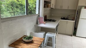 A compact kitchen with a wooden table, white chairs, a gas stove, and a refrigerator. - Jones Beach Haven Studio Escape Kiama Downs opposite patrolled beach and park (KIAMA DOWNS)