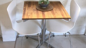 A small, square wooden table with a woven placemat and a potted plant. Two white chairs with metal legs are positioned on either side of the table. - Jones Beach Haven Studio Escape Kiama Downs opposite patrolled beach and park (KIAMA DOWNS)