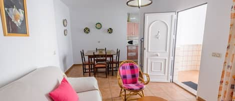 A living room with a sofa, a dining table with chairs, a wicker chair, a small table, and a door leading to a balcony.