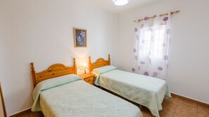 A room with two wooden beds, a nightstand with a lamp, a framed picture on the wall, and a window with floral curtains. - Casas María Carmona - Casa Tierra Níjar by Ruralidays (Almería)