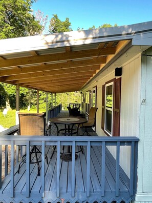 En overdækket terrasse med et bord og stole, gelænder og udsigt til et grønt landskab.
