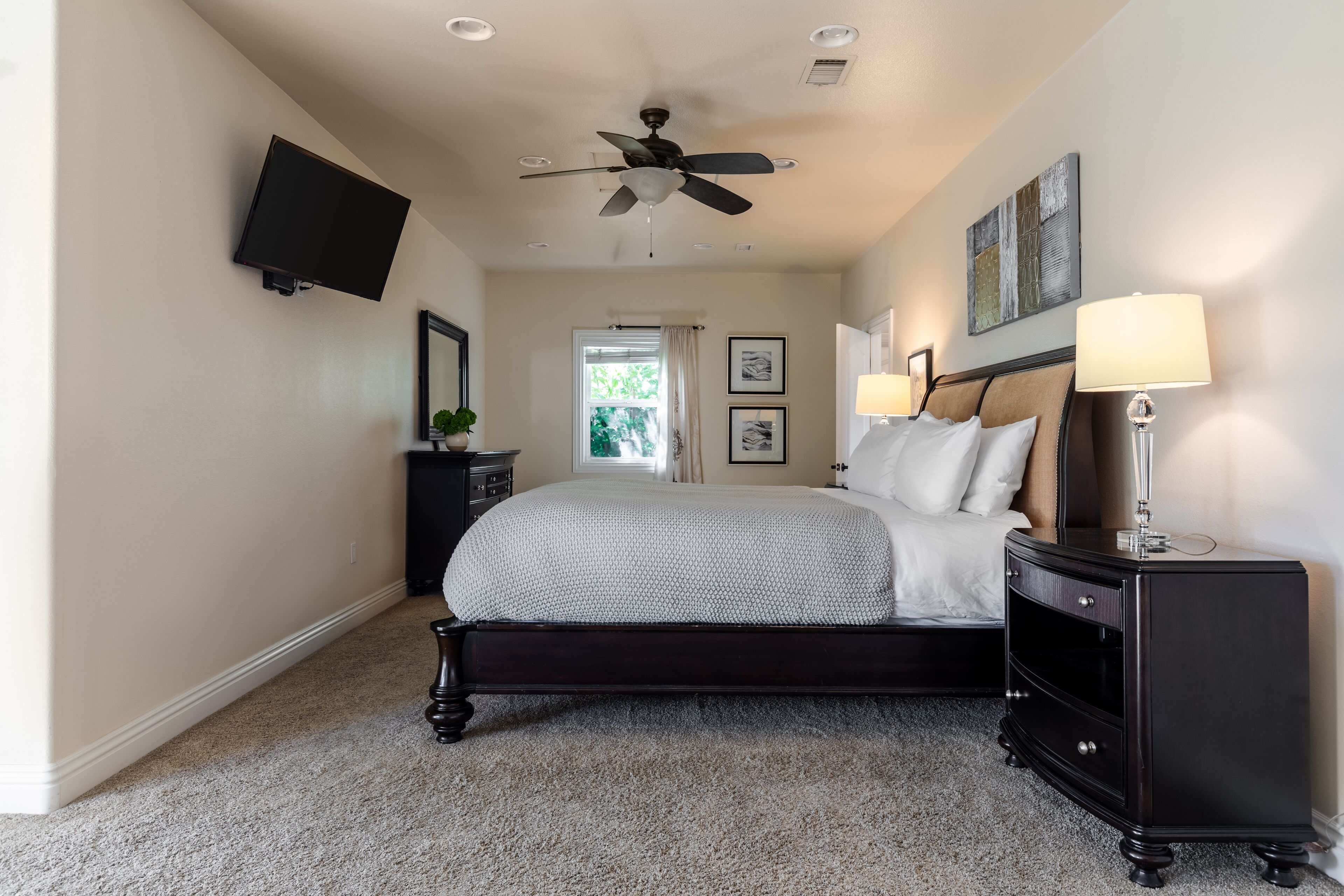 A bedroom with a bed, a nightstand, a TV mounted on the wall, a ceiling fan, and a window with curtains.