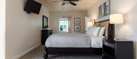 A bedroom with a bed, a nightstand, a TV mounted on the wall, a ceiling fan, and a window with curtains.