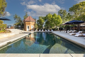 A swimming pool with lounge chairs and umbrellas, surrounded by trees and a small building. - Sleeps 20 | Vineyard Estate w/ Resort Pool, Spa & Game Room | Wilson Creek Manor (Temecula)