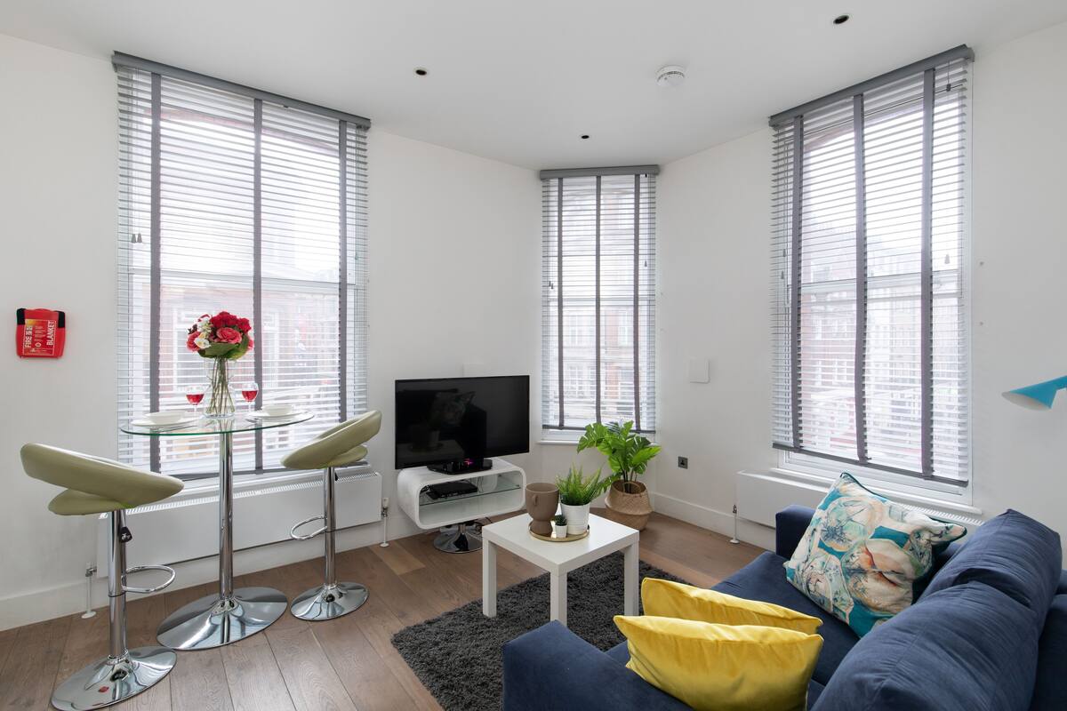 A modern living room with a sofa, a glass table, a TV, and large windows with blinds.
