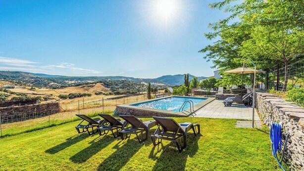 A pool with lounge chairs, a stone wall, and a view of the countryside.
