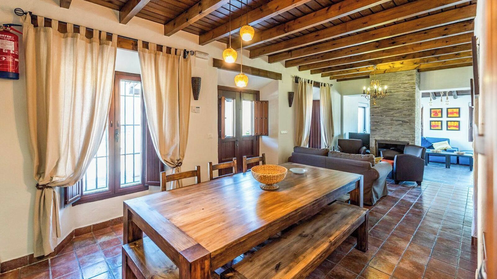 A dining area with a wooden table, chairs, and a bench. There are curtains on the windows, and a fire extinguisher is mounted on the wall.