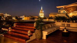 Terraza en la azotea con un sofá, plantas en macetas y faroles. Se observa una torre del reloj al fondo.