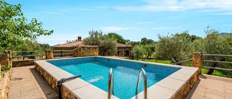 Una piscina rectangular con borde de piedra y escalera metálica, rodeada de muros de piedra y un edificio con techo de piedra al fondo.