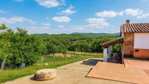 Uma casa rústica com telhado de telhas, um pátio pavimentado e vista para uma paisagem verdejante com colinas ao fundo.