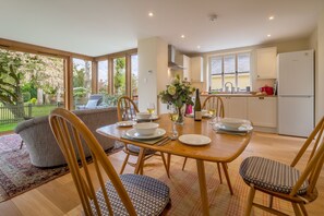 A dining area with a wooden table set for four, surrounded by chairs with patterned cushions. A sofa and a dining table with a vase of flowers are visible in the background.