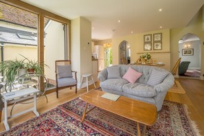 A living room with a grey sofa, patterned cushions, a wooden coffee table, and a red and blue patterned rug. There is a glass table with chairs and a potted plant.
