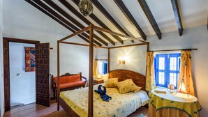 Ein Zimmer mit einem Himmelbett, einer Holzbank, einem kleinen Tisch mit Lampe und einem Fenster mit blauen Vorhängen.
