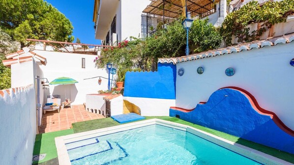 A swimming pool with a blue and white wall, a small patio area with a table and chairs, and a view of a building with a red roof. - Villa Carlitos Comares by Ruralidays (Málaga)
