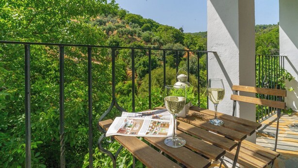 Ein Holztisch mit einer Zeitung, zwei Gläsern Weißwein und einer Bank auf einem Balkon mit Metallgeländer, der auf eine üppige grüne Landschaft blickt.
