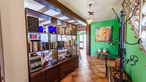 Un pasillo con una estantería de madera repleta de libros y objetos decorativos, una pared verde con un cuadro y un perchero metálico con jarrones.