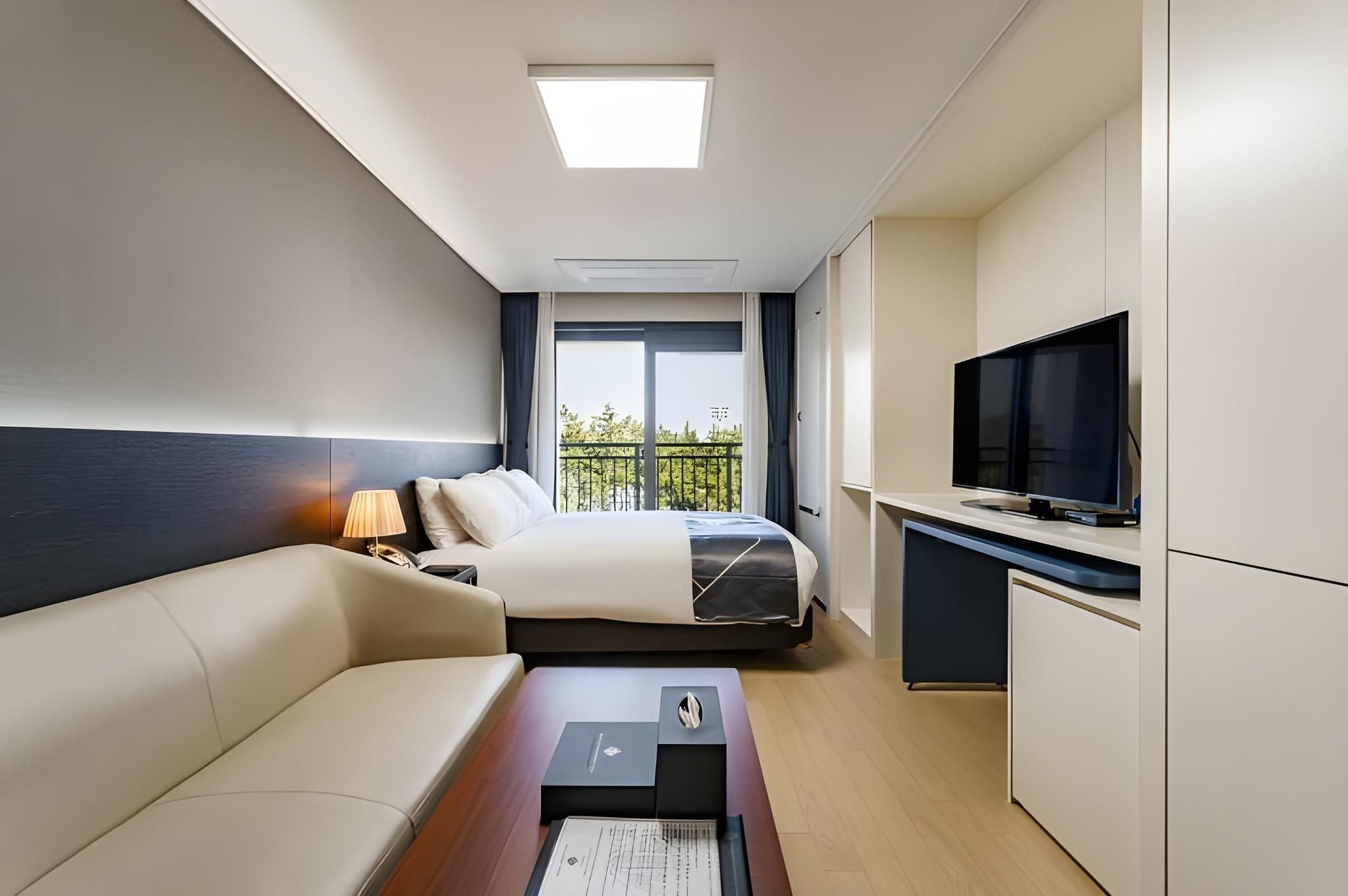 Ein Hotelzimmer mit Bett, Schreibtisch mit Fernseher, einer Sitzecke und einem Fenster mit Blick auf Grünflächen.